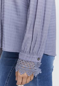 Camicia viola chiaro in tessuto testurizzato, con maniche plissettate e un raffinato bordo in pizzo ai polsini. Presenta un bottone decorativo al polso.