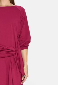 Fioletowa sukienka z rękawami do łokci, detalem w postaci zgromadzonego pasa i wiązaniem z boku. Miękka, gładka tkanina o luźnym kroju i subtelnym połysku.