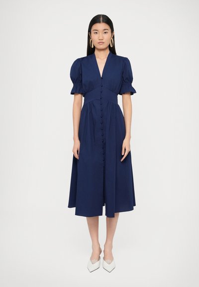 Marineblaues Midikleid mit tiefem V-Ausschnitt, gepufften kurzen Ärmeln und Knopfleiste vorne. Glatte Textur und fließende Silhouette. Kombiniert mit weißen High Heels.