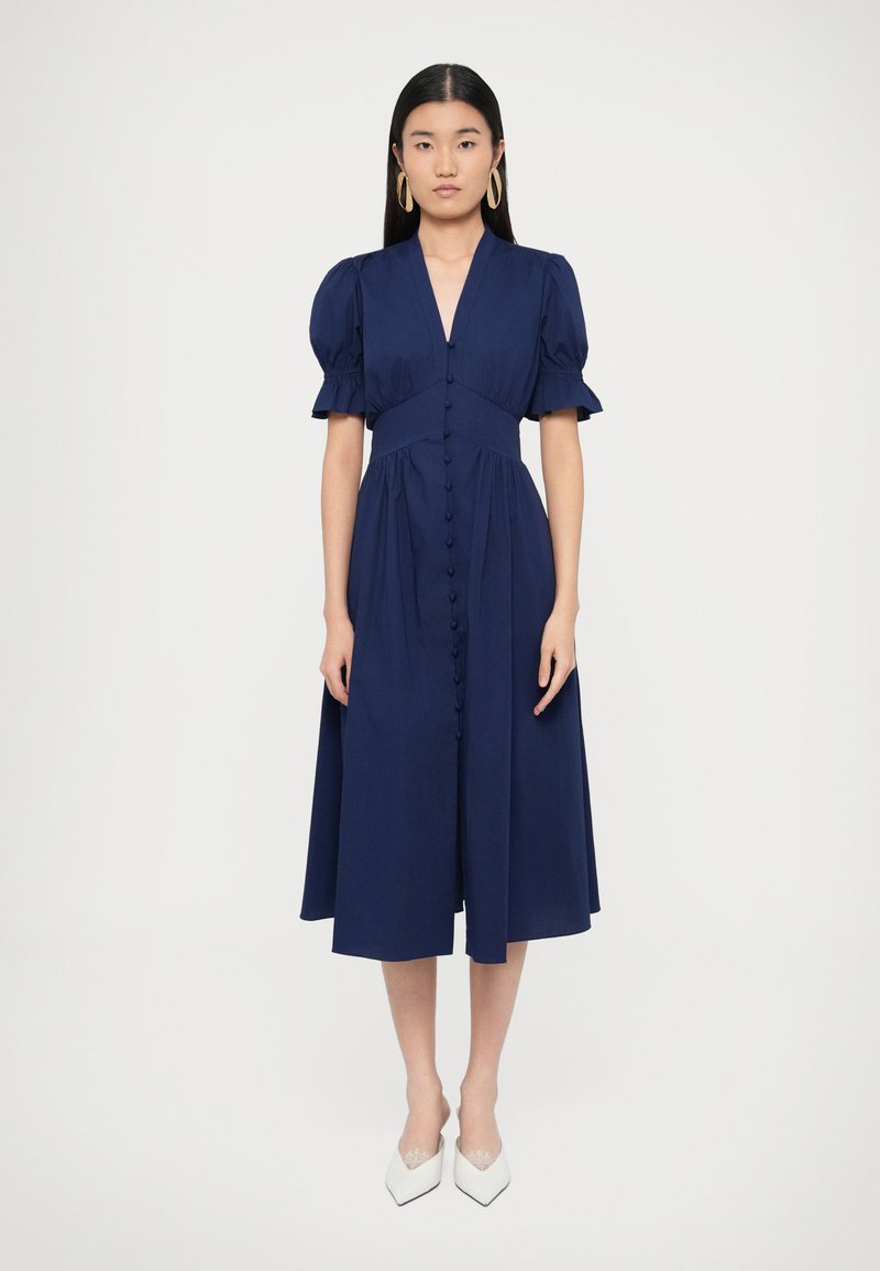 Μίντι φόρεμα σε ναυτικό μπλε με βαθύ V λαιμόκοψη, �φουσκωτά κοντά μανίκια και κουμπιά στο μπροστινό μέρος. Λείο ύφασμα με ρέουσα σιλουέτα. Φορεμένο με λευκές γόβες.