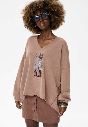 Lichtbruine oversized trui met een V-hals en losse mouwen, voorzien van een kattenafbeelding. Gecombineerd met een bruine jurk met knopen aan de voorkant.