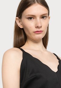 Camisola de cetim preta com decote em ondas, alças finas e textura subtil. Realça as linhas limpas e o tecido suave.