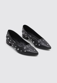 Mocasines de cuero negro con punta afilada, adornados con tachuelas plateadas a lo largo. Tacón bajo y cuadrado. Textura suave con un diseño elegante.