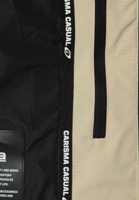 Giacca in tessuto beige e nera con una texture liscia, dotata di una cerniera nera, etichetta con "CARISMA CASUAL" e una tasca laterale.