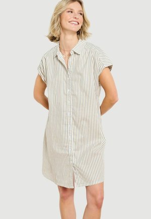 Žena sa usmieva s rukami za chrbtom, oblečená v béžovo-bielej krátkorukávovej šaty s vertikálnymi pruhmi a gombíkmi na svetlom pozadí.