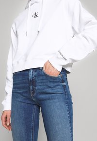 Witte hoodie met een logo op de borst en merknamen op de mouw, gecombineerd met blauwe denim jeans met een aangesloten pasvorm en lichte veroudering.