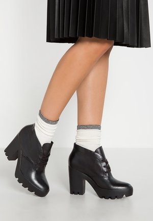 Jambes avec des chaussettes blanches aux chevilles et des chaussures noires épaisses à talons hauts lacées, avec une jupe noire plissée visible au-dessus des genoux.