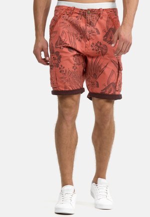Shorts made of orange fabric featuring dark tropical floral patterns, with side pockets and rolled hems. Worn with white sneakers.