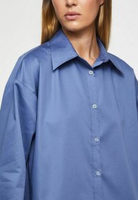 Ljusblå skjorta med knappar, styv krage och vida ärmar. Mjuk tygstruktur med subtil glans och sju knappar framtill.