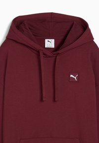 Burgundi kapuutsärk, mis on valmistatud pehmest kangast. Omab eesmist känguru taskut, reguleeritavat kapuutsi ja valget Puma logo plaastrit rinnal.