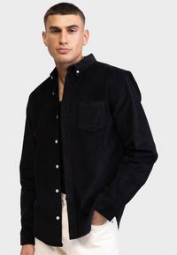 Camicia in corduroy nera con colletto button-down, taschino singolo sul petto e bottoni bianchi. Il tessuto texturizzato aggiunge profondità al design.