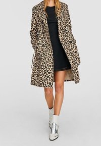 Trench en estampado de leopardo en beige y negro, diseño de apertura frontal, manga larga, combinado con un vestido negro y botines plateados.