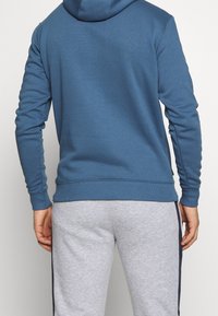 Felpa blu con cappuccio, con polsini e orlo a costine, abbinata a pantaloni sportivi grigi chiari con una striscia laterale. Tessuto morbido, vestibilità rilassata.