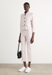 Roze blazer met grote kraag, zwarte knopen en voorzakken, gecombineerd met bijpassende aansluitende broek. Witte sneakers en een zwarte schoudertas.