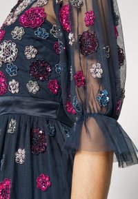 Marinblå tulleklänning prydd med flerfärgade paljettblomm önster. Har puffärmar med volangkant och en rynkad midja.