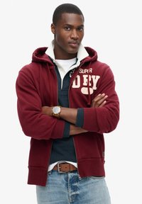 Felpa con zip bordeaux con tasca a marsupio e cappuccio con coulisse, indossata sopra un polo blu navy, abbinata a jeans e una cintura marrone.
