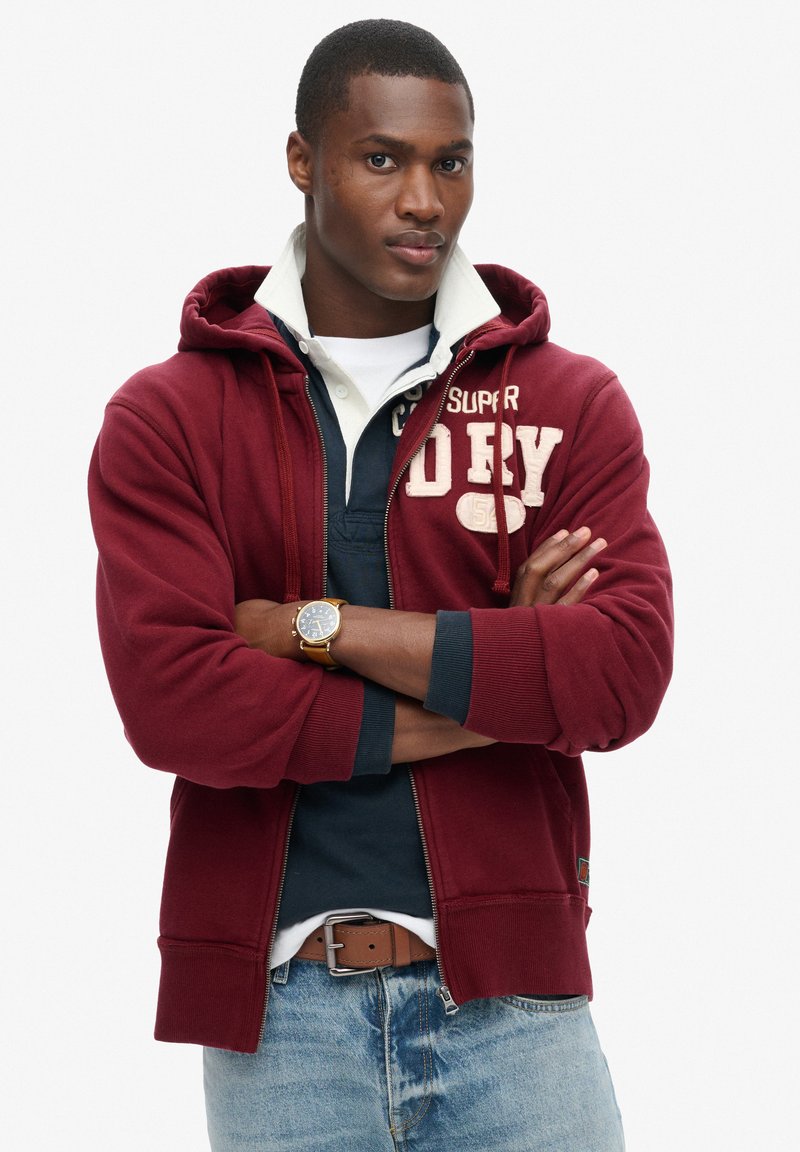 Felpa con zip bordeaux con tasca a marsupio e cappuccio con coulisse, indossata sopra un polo blu navy, abbinata a jeans e una cintura marrone.