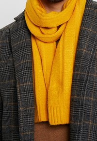 Écharpe jaune en tricot portée sur une chemise marron, associée à un blazer à carreaux gris foncé présentant un tissu texturé et une finition lisse.