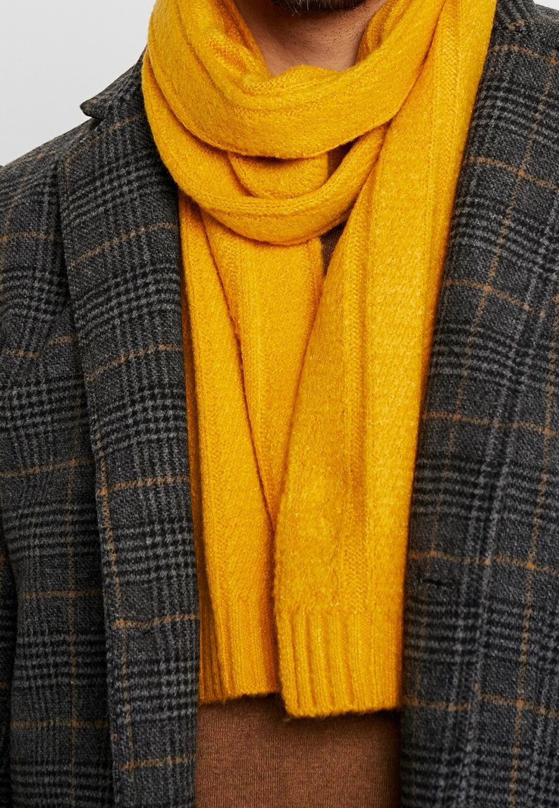 Écharpe jaune en tricot portée sur une chemise marron, associée à un blazer à carreaux gris foncé présentant un tissu texturé et une finition lisse.