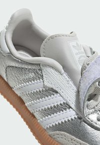 Close-up af en sølvfarvet Adidas Samba sneaker med hvide striber, beige tungetiket, beige snørebånd og sål i gummigummi.