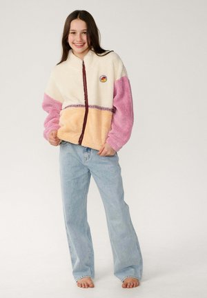 Lächelndes Mädchen steht barfuß und trägt eine Farbblock-Fleecejacke in Creme, Pink und Pfirsich sowie hellblaue Jeans vor einfachem Hintergrund.