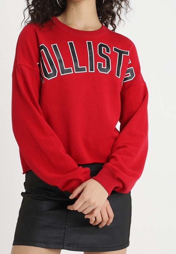 Felpa rossa con grande scritta nera "HOLLISTER" sulla parte anteriore. Presenta spalle scese e maniche lunghe. Abbinata a una gonna di pelle nera.