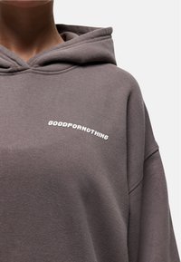 Persoon in een donkergrijze hoodie met witte tekst "GOODFORNOTHING" op de linkerkant van de borst, zichtbaar van de nek tot halverwege de torso.