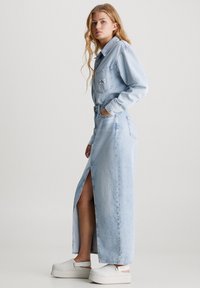 Mujer de pie de lado, vestida con una camisa de mezclilla azul claro y una falda larga de mezclilla con una abertura frontal, acompañada de zuecos de plataforma blancos.