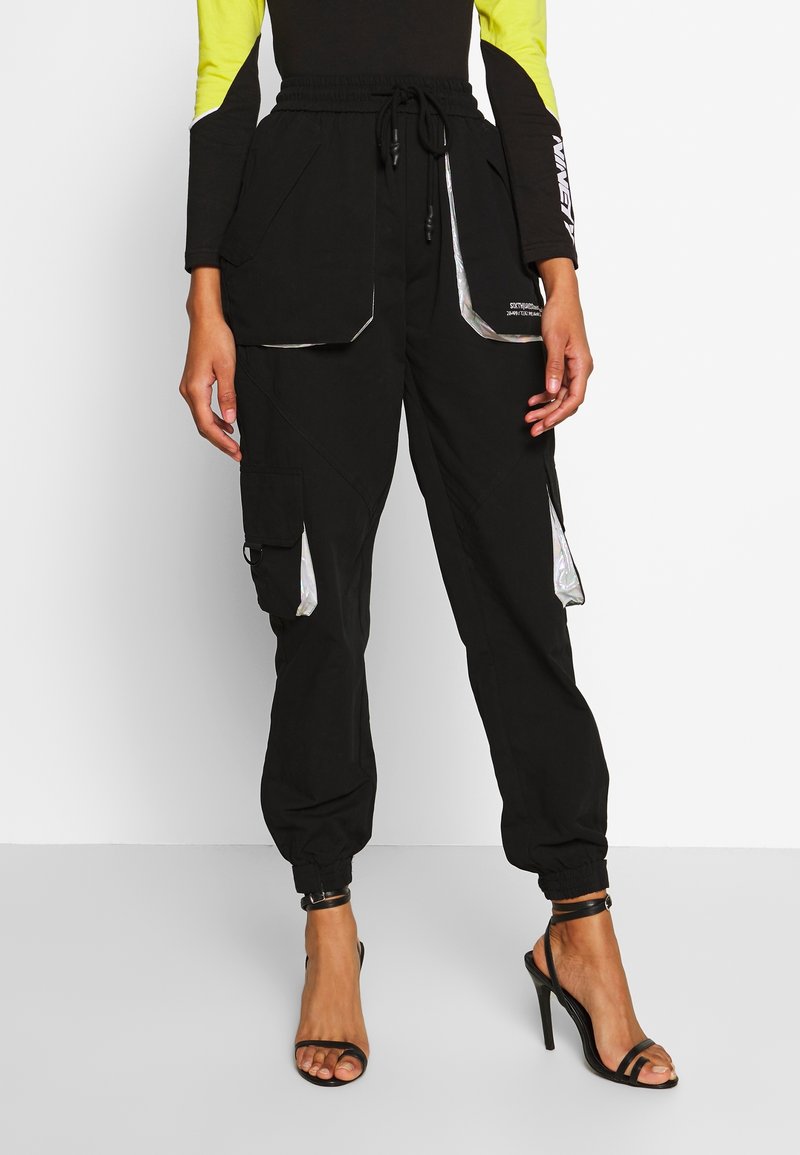 Zwarte cargobroek met reflecterende accenten, elastische tailleband, meerdere zakken en taps toelopende boorden, gecombineerd met zwarte sandaaltjes met bandjes.