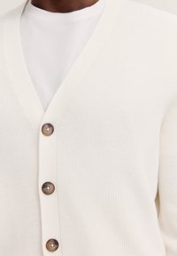 Cremefarbener gestrickterCardigan mit V-Ausschnitt, fünf braunen Knöpfen und einem gerippten Muster. Getragen über einem weißen T-Shirt.