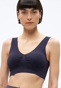 Moteris su trumpais rudais plaukais, vilkinti tamsiai mėlyną vientisą sportinę liemenėlę, stovinti priešais paprastą šviesų foną.