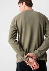 Langsleeve shirt in olijfgroen, gemaakt van textuurstof met ribbels. Heeft een ronde halslijn en aangesloten manchetten. Achteraan is een ontspannen pasvorm zichtbaar.