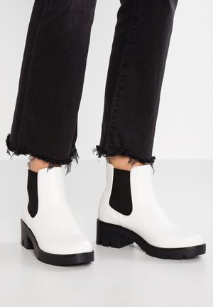 Botas de tobillo de cuero blanco con paneles laterales de elastano negro, suelas gruesas negras y un acabado liso, combinadas con jeans negros deshilachados.
