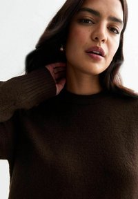 Kvinna med mörkt hår som bär en brun långärmad tröja, rör vid sin hals, med neutral uttryck och guldörhängen i form av ringar.