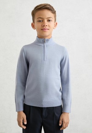 Pull bleu clair à manches longues avec fermeture éclair, col montant, texture lisse et poignets côtelés, associé à un pantalon foncé.