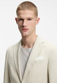 Hombre rubio con cabello corto que lleva un blazer crema texturizado sobre una camiseta gris clara, posando frente a un fondo blanco liso.