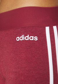 Burgundin urheiluleggarit, joissa on leveä vyötärönauha, valkoinen Adidas-logo ja kolme valkoista sivuhihnaa. Pehmeä, joustava kangas, jossa on sileä pinta.