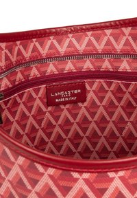 Intérieur en tissu rouge d'un sac avec un motif géométrique en triangles. L'étiquette indique "LANCASTER PARIS, FABRIQUÉ EN ITALIE," avec une poche zippée.
