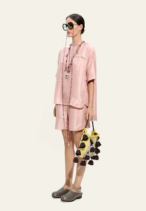 Kobieta w różowej satynowej sukience, nosząca duże okrągłe okulary przeciwsłoneczne, długie koralikowe naszyjniki, szare chodaki i trzymająca żółtą torbę z czarnymi chwostami.