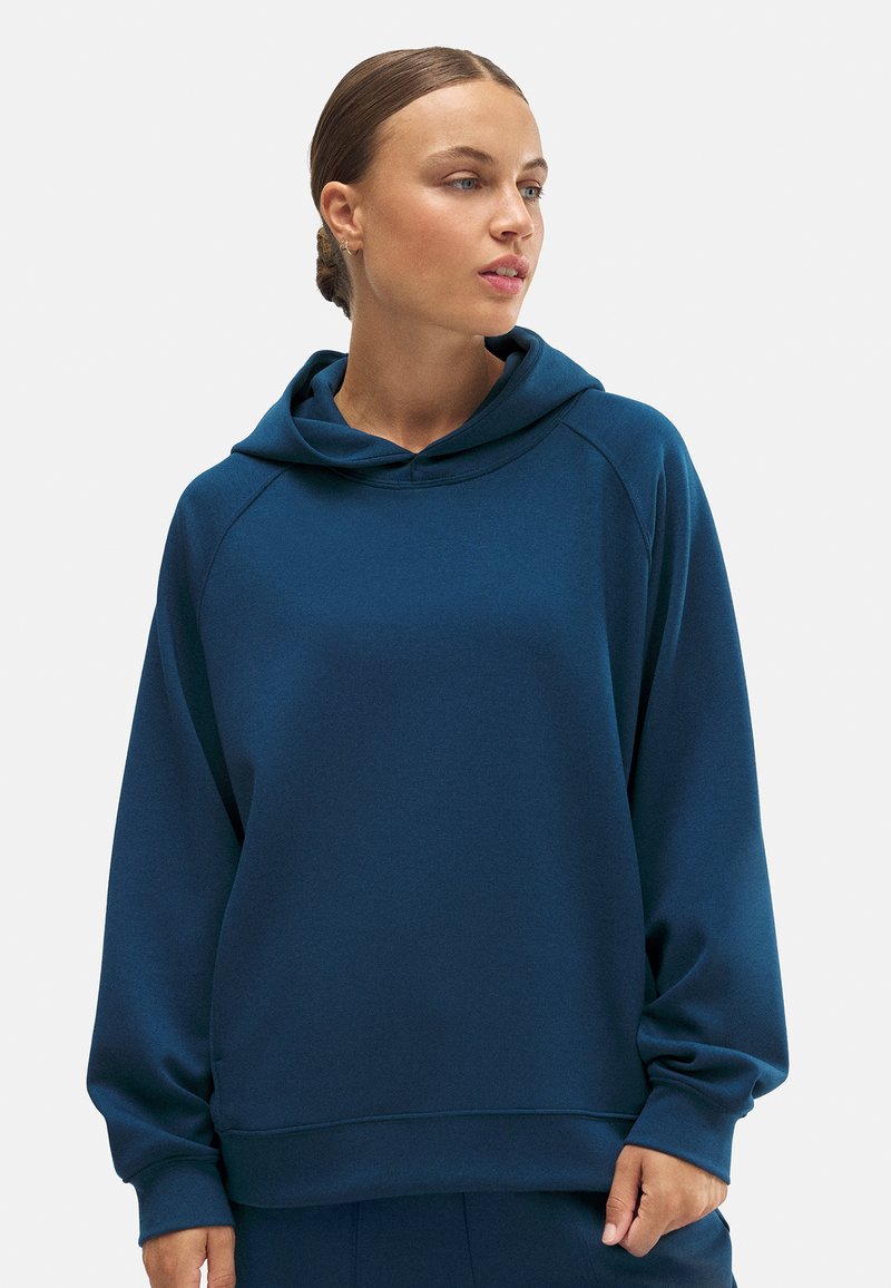 Sudadera azul con capucha que presenta un corte relajado, mangas raglán y una textura suave. La tela es lisa, con costuras y detalles mínimos.