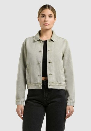 Fiatal nő, aki világosszürke, gombos farmerdzsekit visel fekete, rövid fazonú felső és fekete, magas derekú farmernadrág fölött, egyszínű háttér előtt áll.