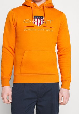 Mand iført en orange hoodie med GANT-logo, skjoldemblem og teksten "American Sportswear", kombineret med mørkeblå bukser.