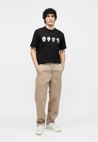 Νέος άνδρας όρθιος με τα χέρια στις τσέπες, φοράει μαύρο μπλουζάκι με τέσσερα πολύχρωμα σχέδια κρανίων, μπεζ παντελόνι και λευκά αθλητικά παπούτσια σε απλό φόντο.