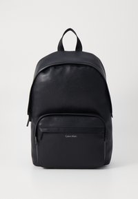 Černý batoh vyrobený z hladkého syntetického materiálu, s kulatým vrškem, přední kapsou na zip a stříbrným logem Calvin Klein.