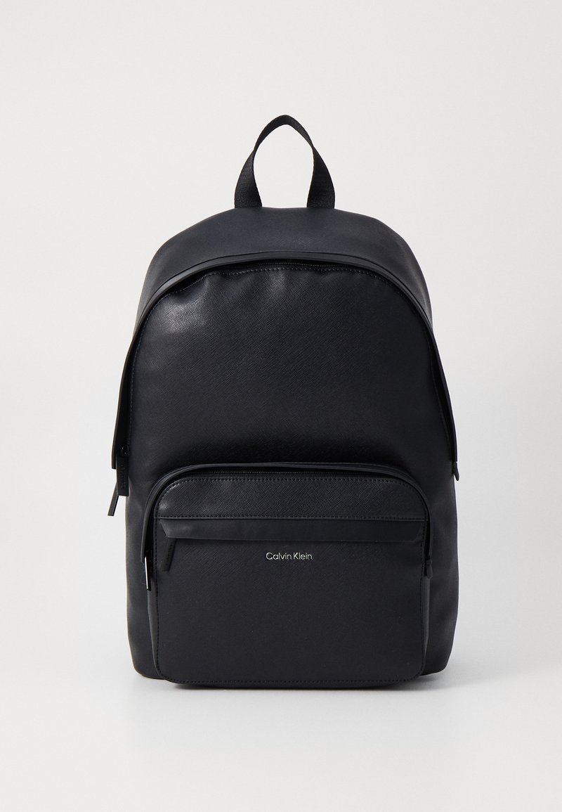 Black backpack made from smooth synthetic material, featuring a rounded top, front zip pocket, and Calvin Klein logo in silver.