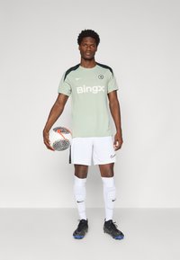 Maillot de football Nike vert et noir avec le logo "Bingx", shorts blancs, chaussures de football noires, et un ballon à motif tenu devant.