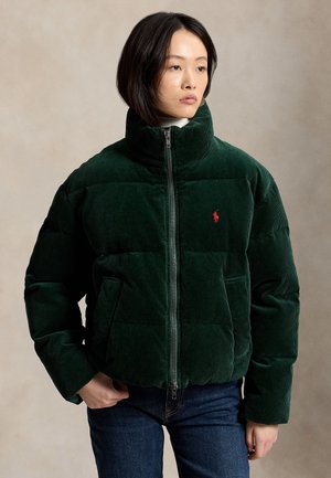 Donna che indossa una giacca imbottita in velluto a coste verde scuro con collo alto e logo rosso, abbinata a jeans blu scuro, su uno sfondo neutro.