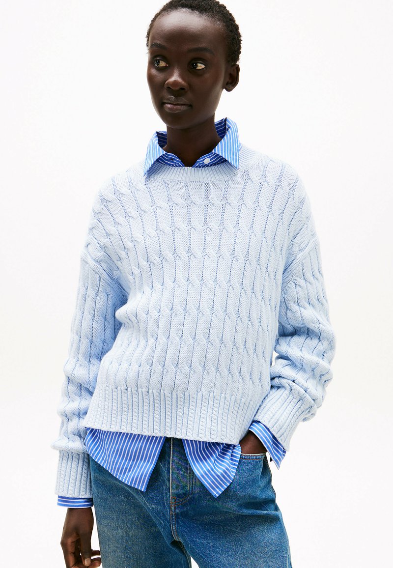 Maglione lavorato a maglia di colore azzurro chiaro con motivo testurizzato, polsini e orlo a coste, indossato sopra una camicia a righe blu e bianche, abbinato a jeans in denim.