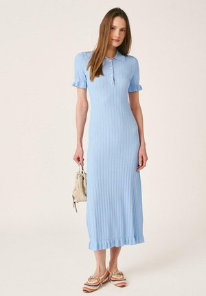 Mujer con un vestido midi azul claro acanalado con mangas y dobladillo con volantes, sosteniendo un bolso tejido beige y llevando zapatos planos de rayas multicolor.