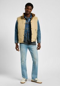 Pufferväst i beige med en kordfluwelekant över en jeansjacka. Ljusblå jeans och krämfärgade sneakers med svarta ränder. En enfärgad bakgrund.